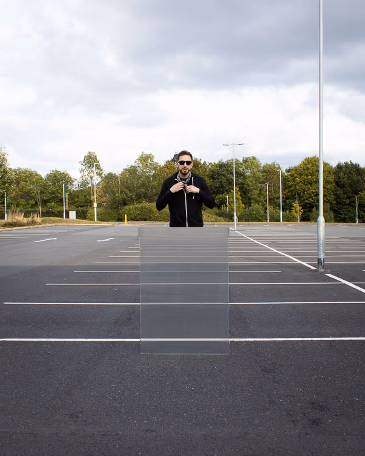 An individual, dressed in a black jacket and sunglasses, is standing in the center of an expansive, vacant parking lot, which is bordered by white lines and flanked by trees and a cloudy sky in the background.