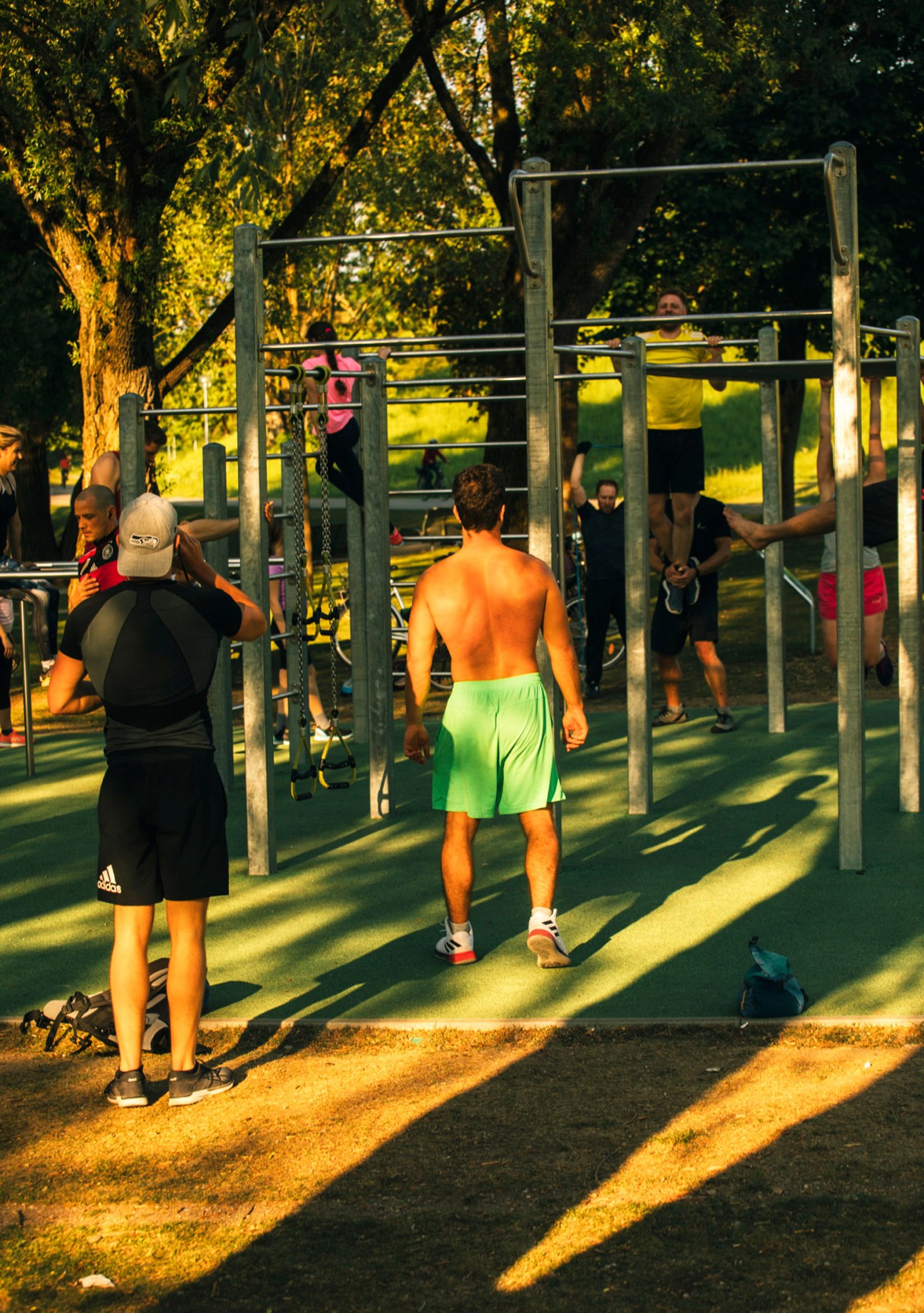 People exercising at outdoor fitness equipment under trees in a sunny park setting.