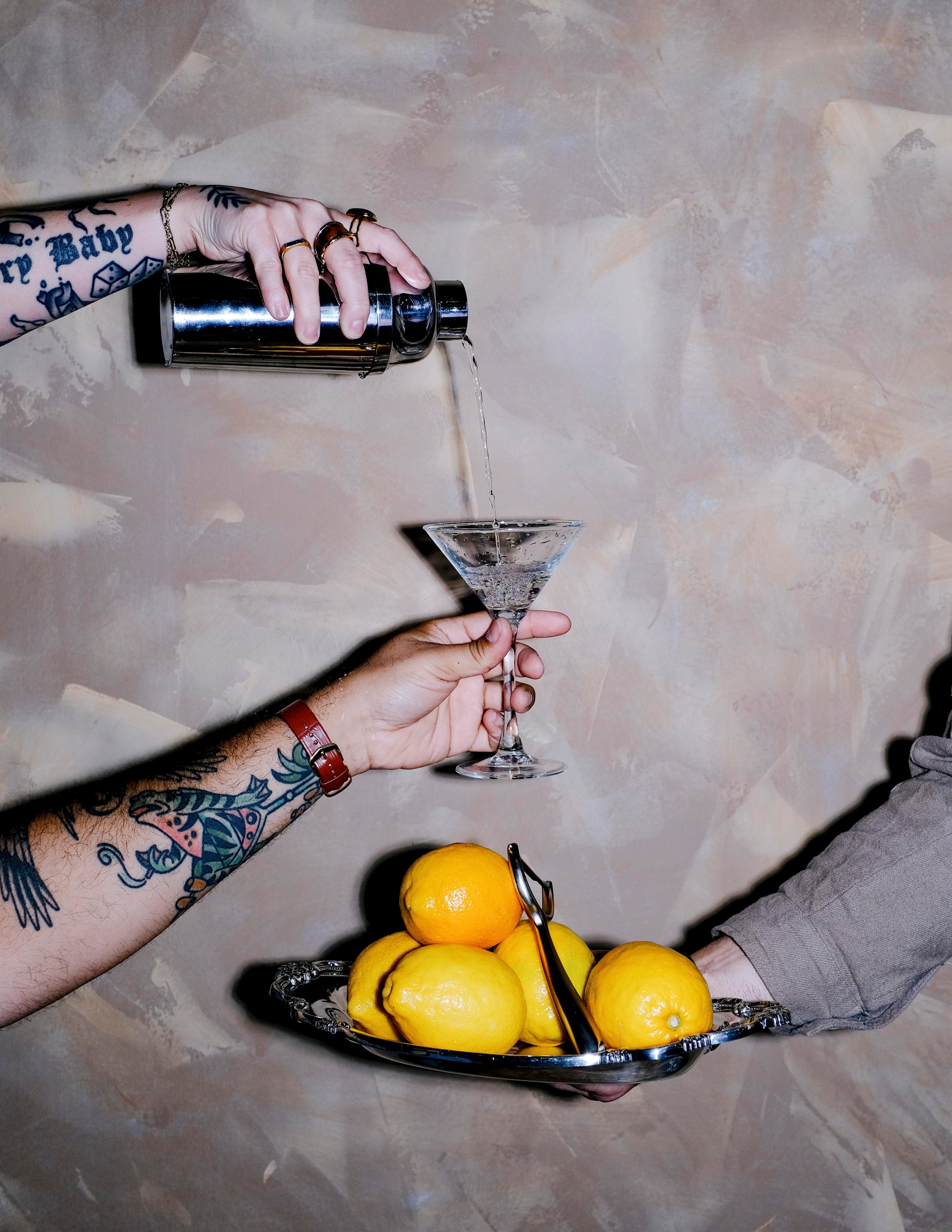 Someone holds a silver dish with citrus while another hand holds out a martini glass as someone pours from a cocktail shaker.