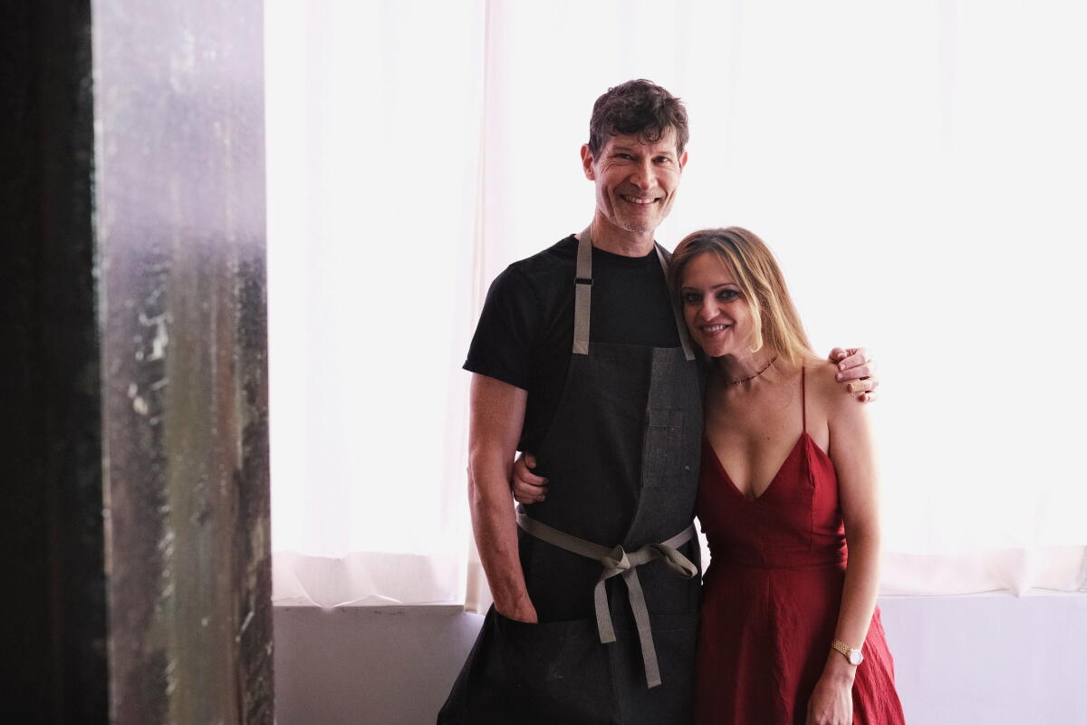 Husband and wife Daniel Patterson and Sarah Lewitinn stand in front of a light purple curtain at Jacaranda