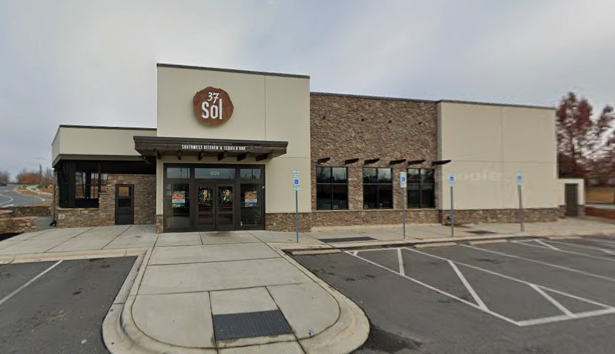 Exterior of “37 Sól Southwest Kitchen & Tequila Bar,” a modern modern commercial building with light beige siding and stone masonry sections. A round wooden-style sign with the logo “37 Sól” hangs above the main entryway overhang, which displays the tagline “Southwest Kitchen & Tequila Bar” in white text. An empty parking lot with designated accessible parking stalls sits in front of the building under an overcast sky.