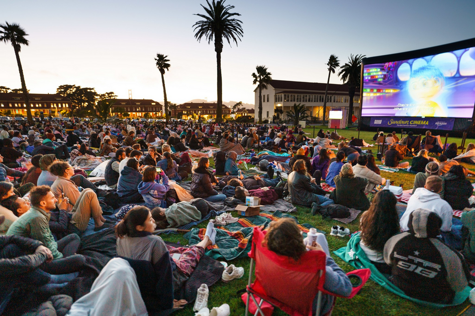 The best events in SF this week, from Sundown Cinema to Restaurant Week A large crowd sits on blankets and chairs on grass, watching an animated movie at an outdoor cinema during sunset, with palm trees and buildings in the background.