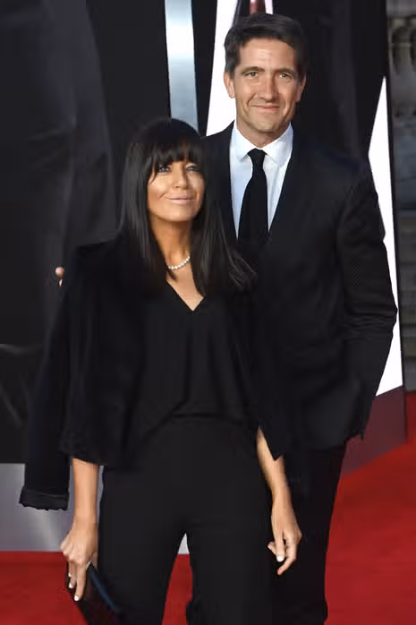 An adult man and woman dressed in formal attire, smiling and standing next to each other against a dark background.
