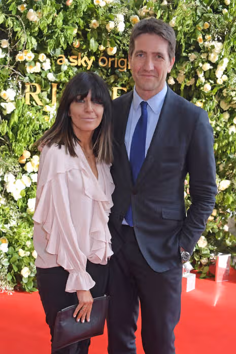 An adult male and an adult female are standing next to each other and smiling for a photo. They are both dressed formally, with the male wearing a suit and tie and the female in a white blouse. The background features a green floral wall with yellow flowers.