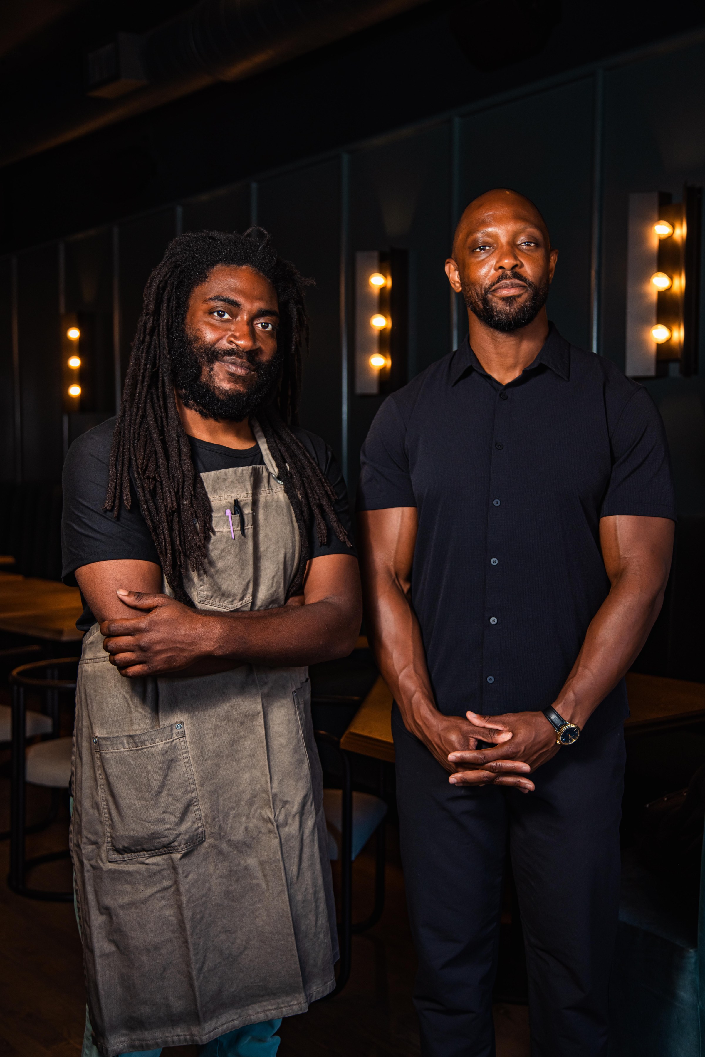 A man with dreadlocks and an apron stands next to a man with his hands folded in front of him.
